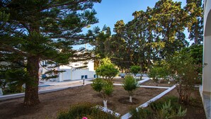 A landscaped garden with various trees and shrubs, a paved pathway, and a white building in the background. - Irene Hotel (Leros)