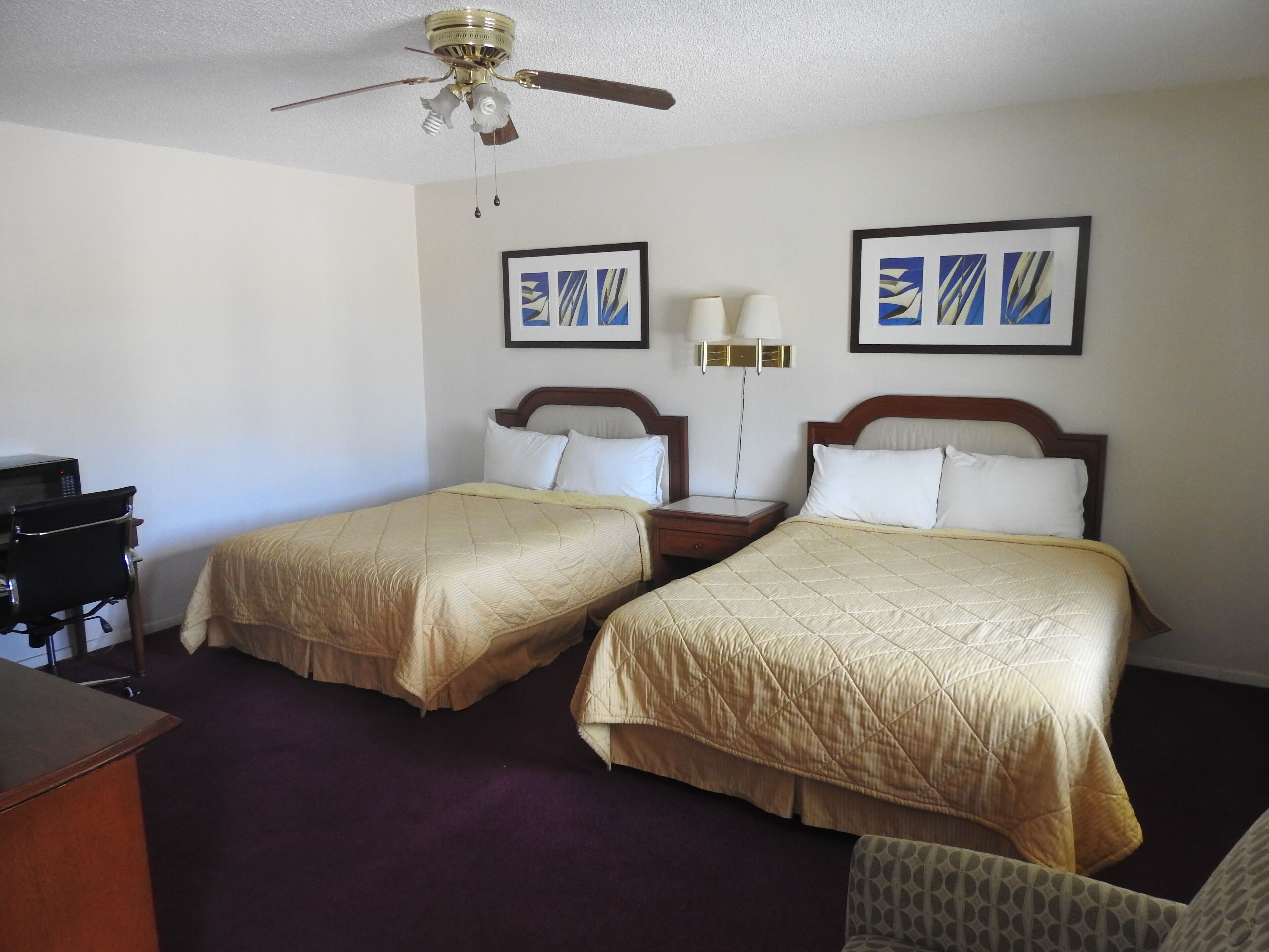 A hotel room with two beds, a desk, a chair, a ceiling fan, and artwork on the walls.
