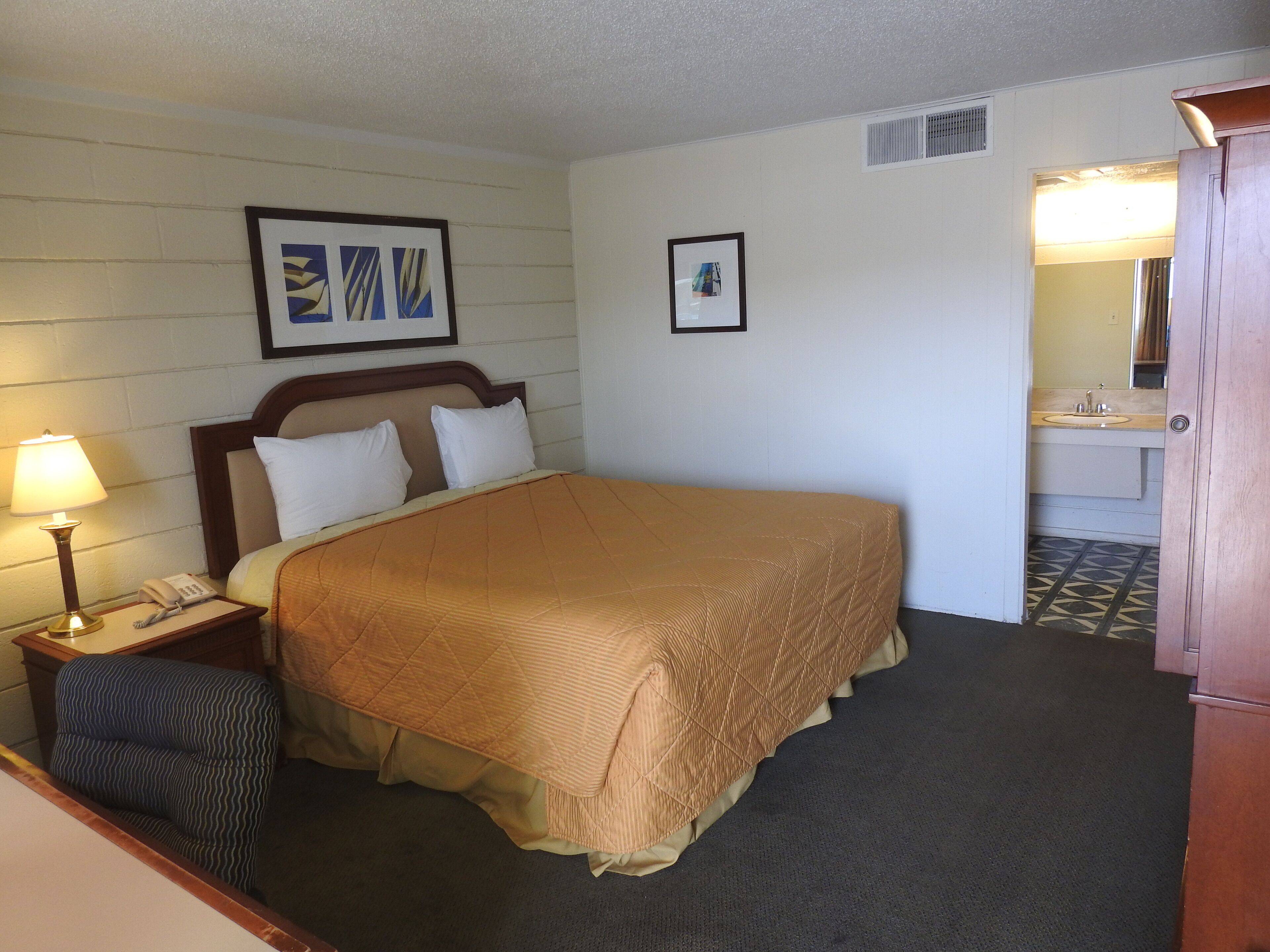 A hotel room with a bed, a nightstand with a lamp, a chair, a framed picture on the wall, and a bathroom visible through an open door.