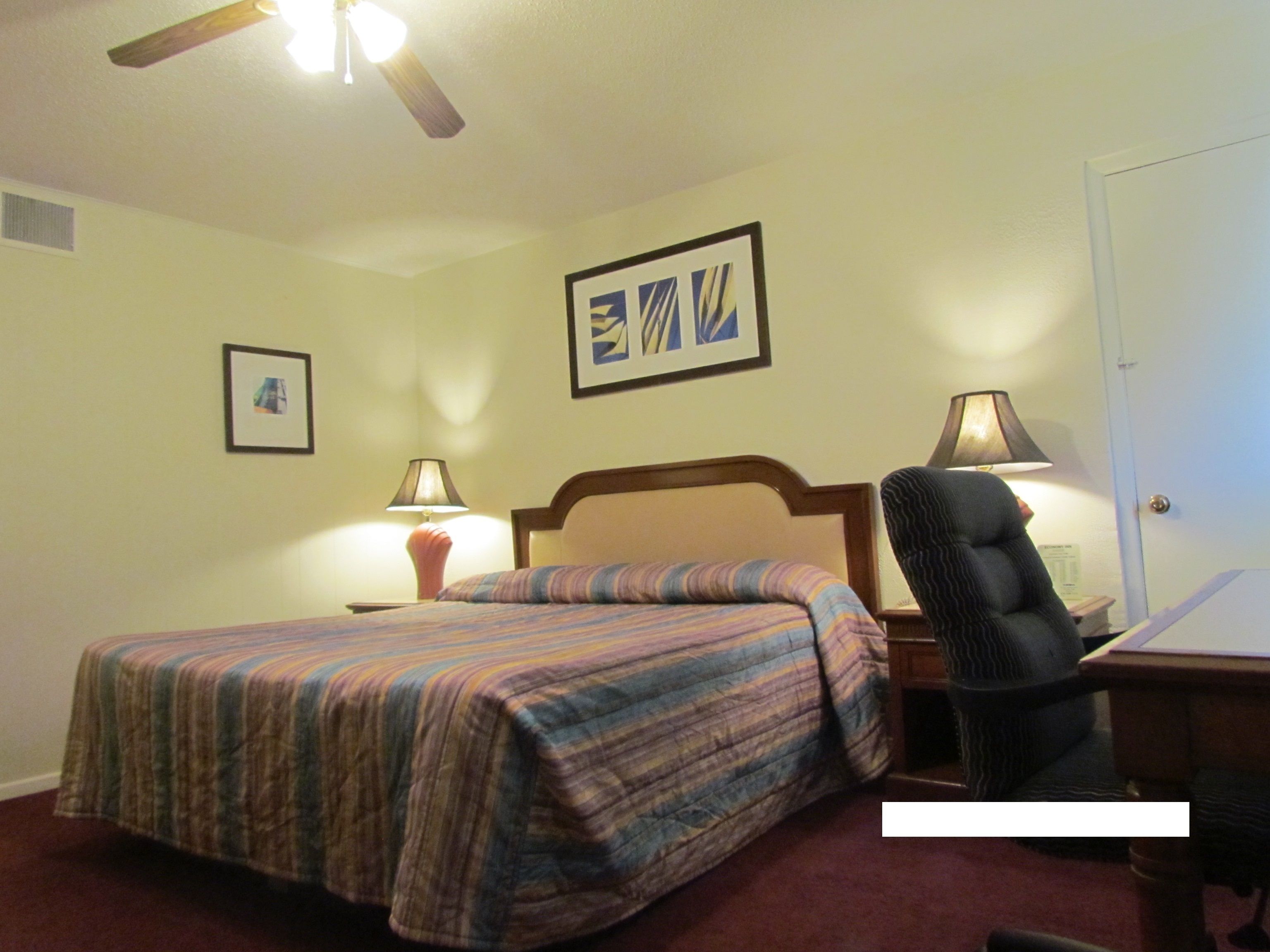 A hotel room with a bed, a chair, two lamps, a ceiling fan, and two framed pictures on the wall.