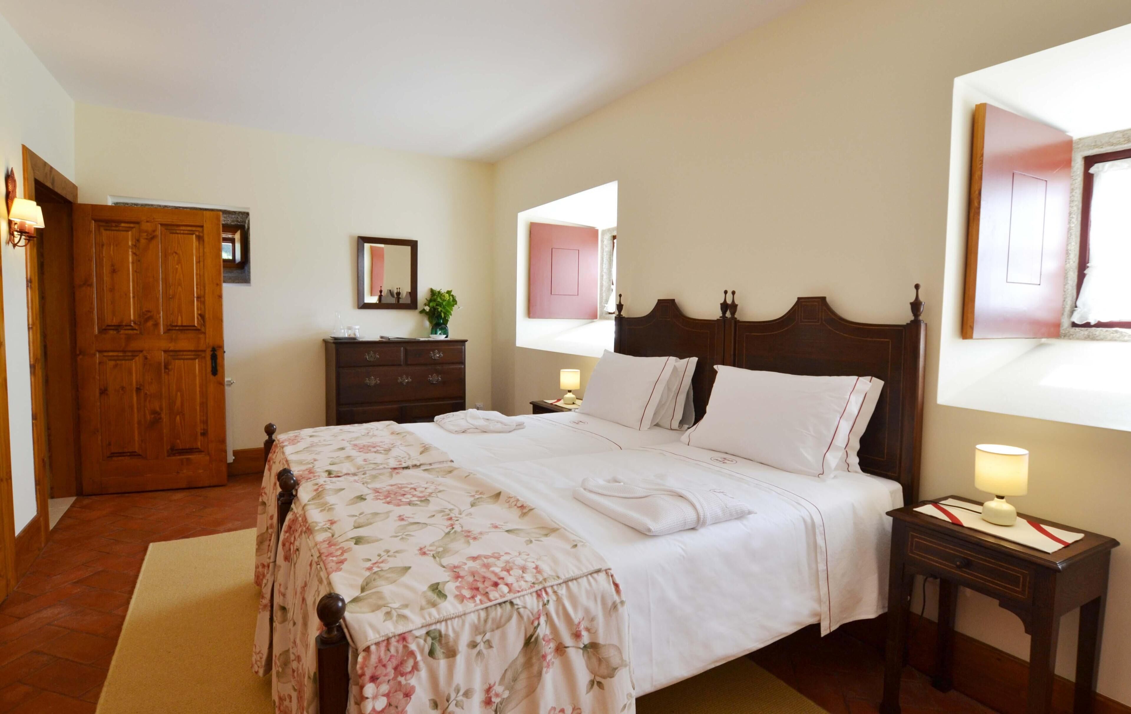 A neatly made bed with a floral bedspread, a wooden wardrobe, a nightstand with a lamp, and a mirror on the wall.