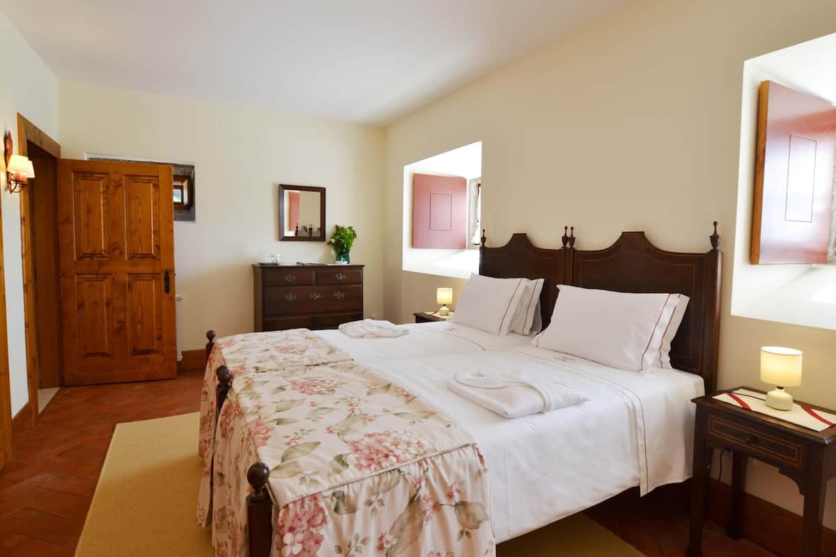 Een net opgemaakt bed met een bloemenbedekking, een houten garderobe, een nachtkastje met een lamp en een spiegel aan de muur.