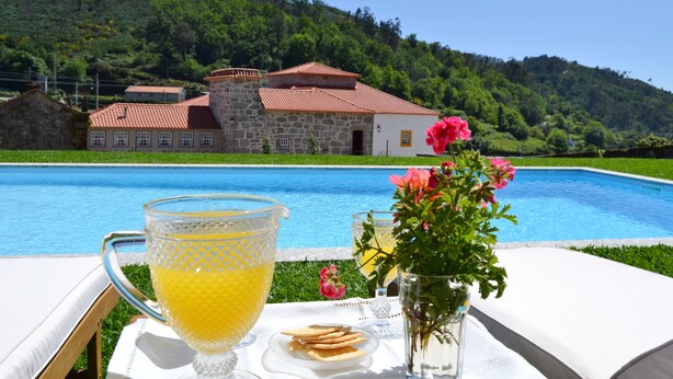 Une scène au bord de la piscine, avec une vue dégagée sur un bâtiment en pierre doté d’un toit rouge, une carafe en verre remplie de jus d’orange et un vase avec des fleurs roses.