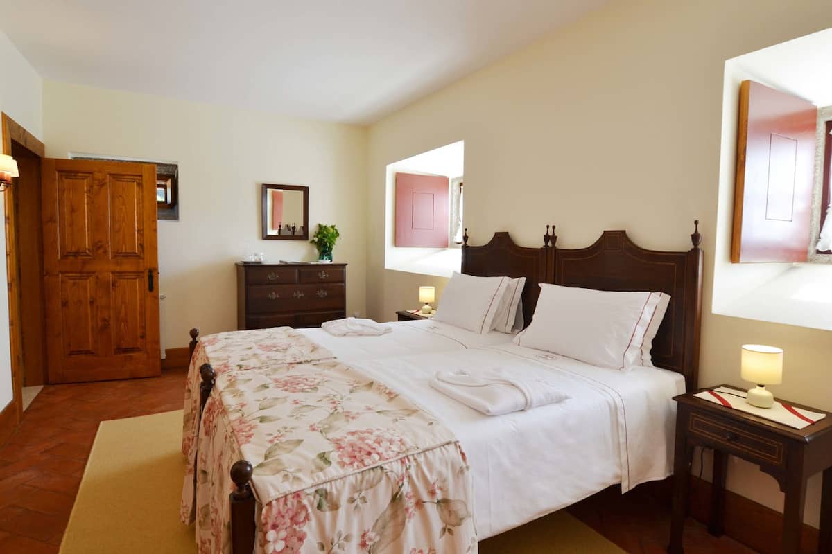 Een net opgemaakt bed met een bloemenbedekking, een houten garderobe, een nachtkastje met een lamp en een spiegel aan de muur.