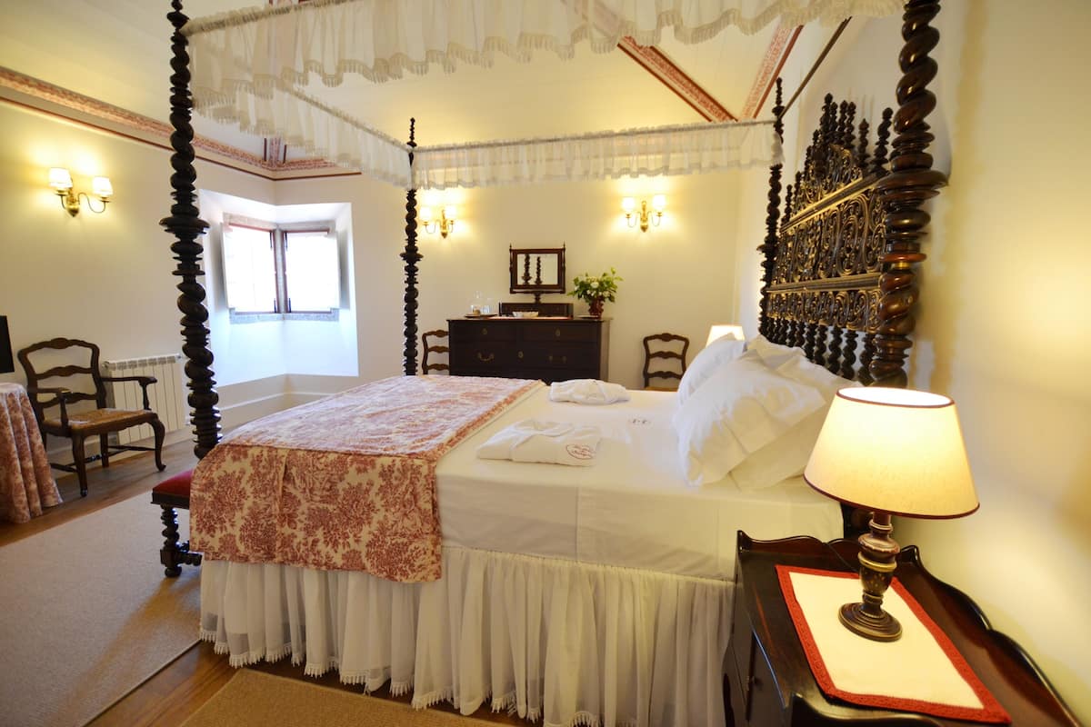 Een hemelbed met een baldakijn, een nachtkastje met een lamp, een dressoir en een stoel.