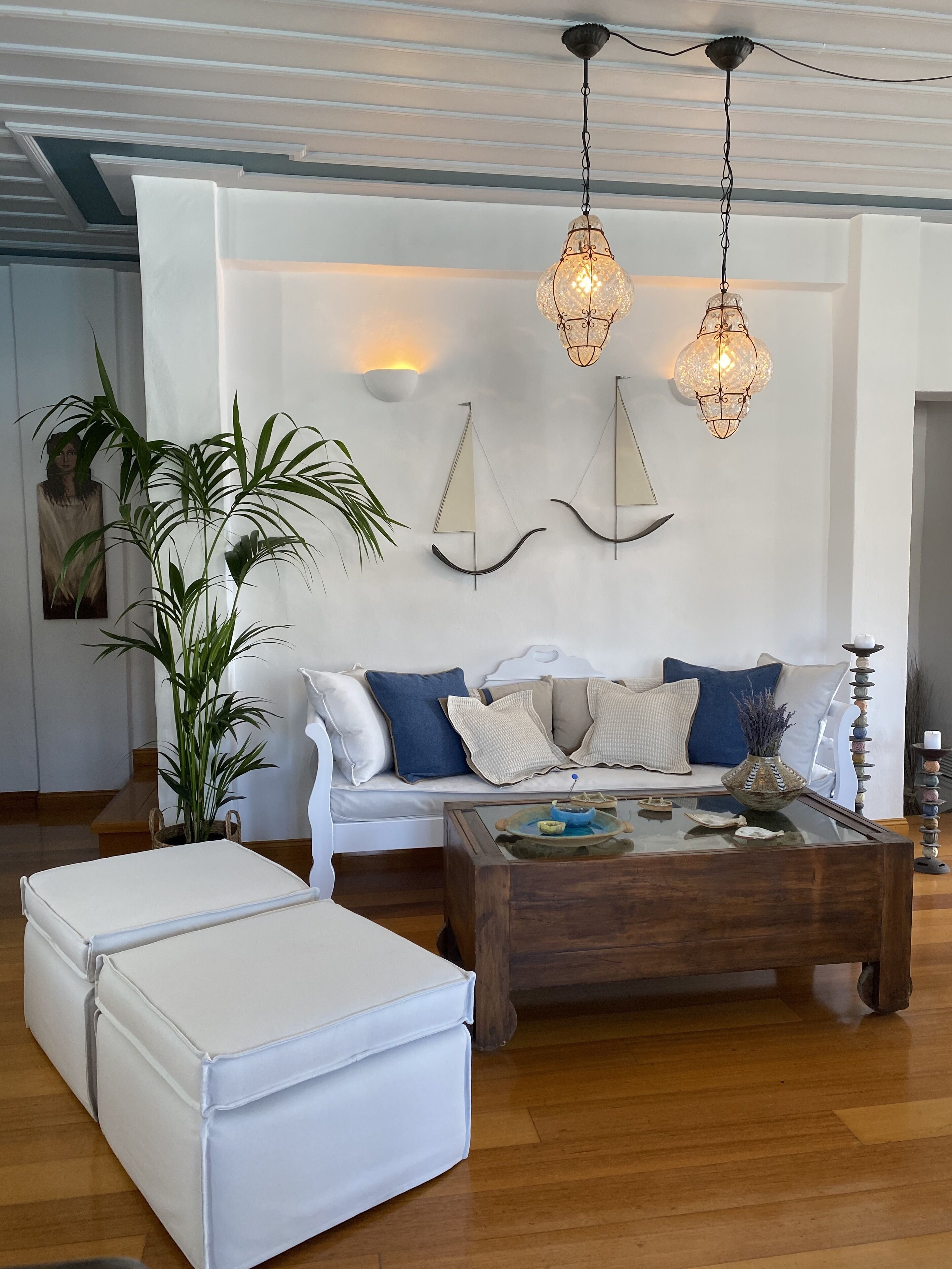 A modern living room with a white sofa, a wooden coffee table, and a white ottoman. There are two hanging pendant lights, a wall-mounted shelf with decorative items, and a potted plant.