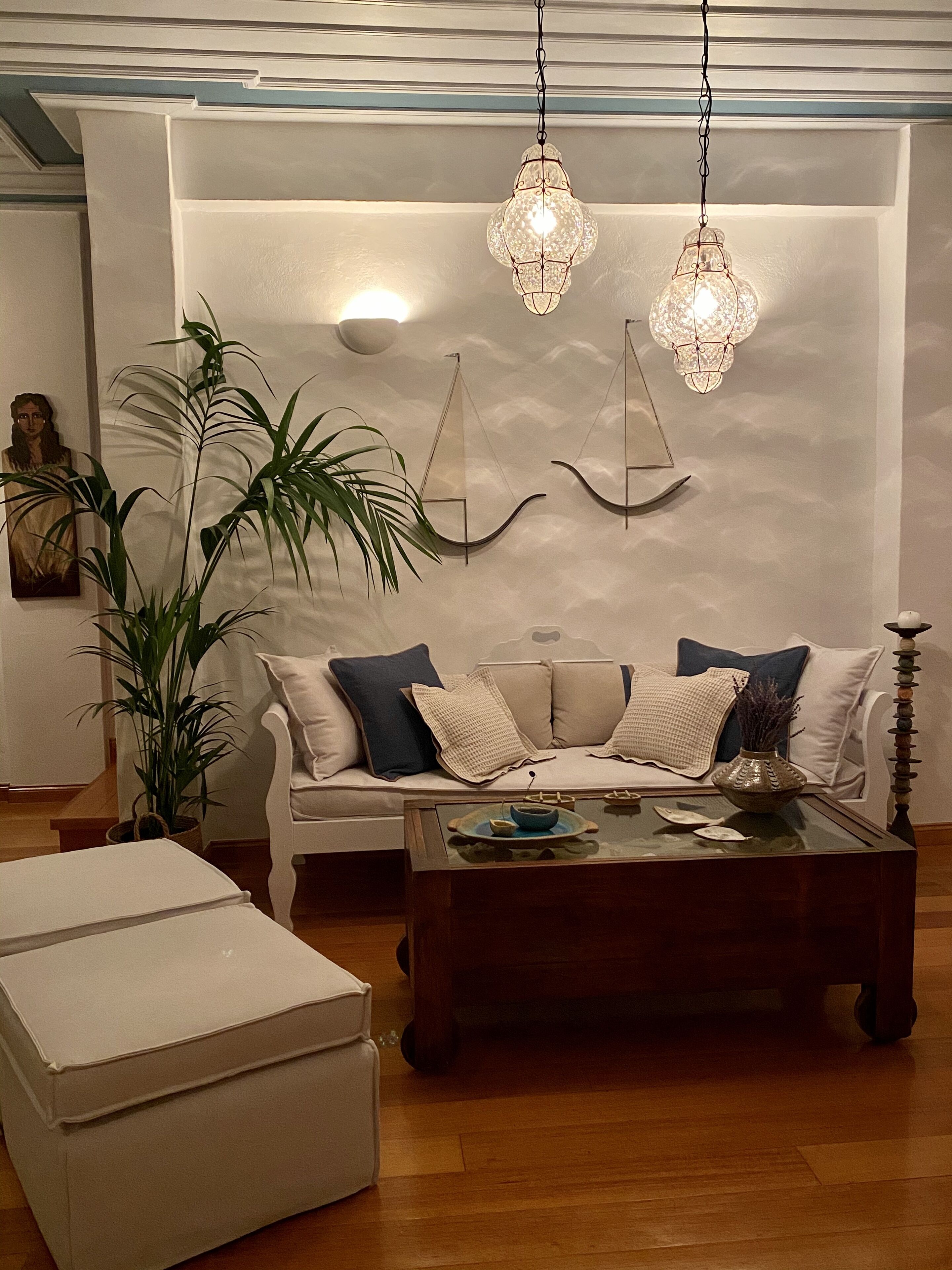 A modern living room with a white sofa, a glass coffee table, and a white ottoman. There are two hanging crystal chandeliers, a large potted plant, and a wall-mounted sculpture.