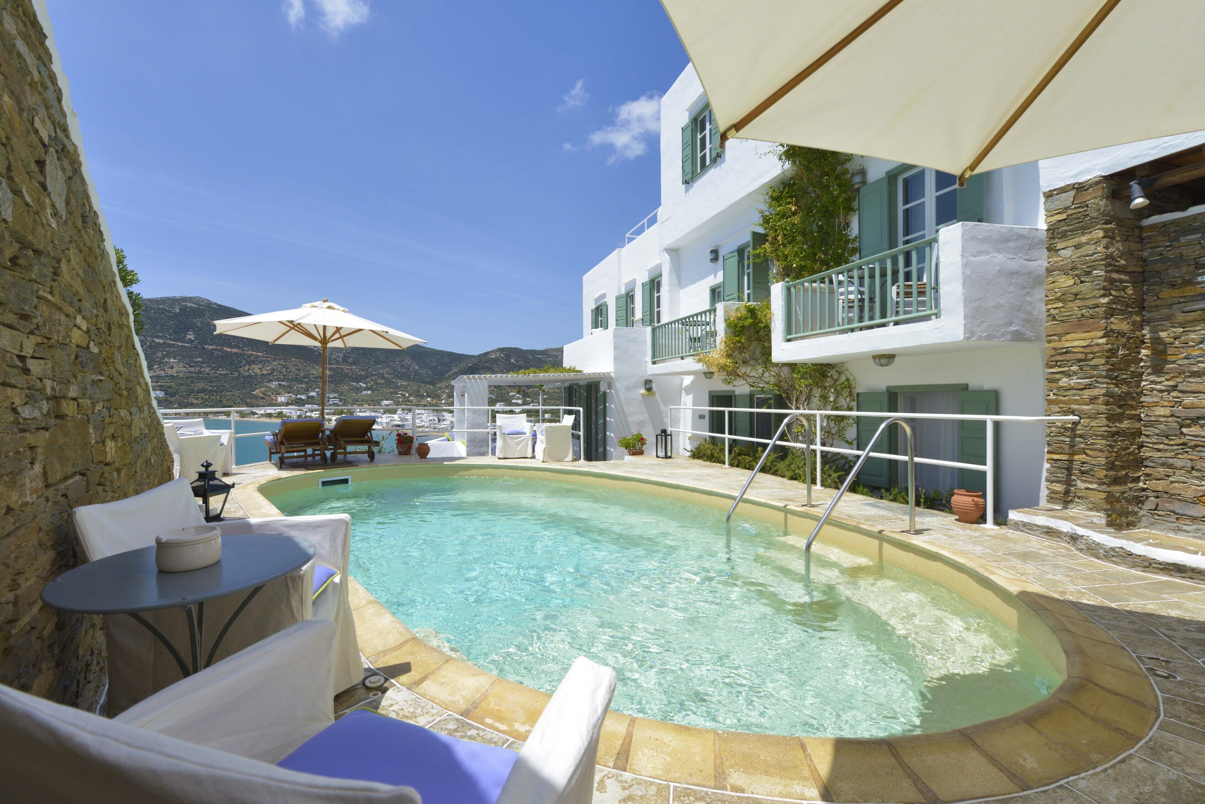 Uma área de piscina com espreguiçadeiras e guarda-sóis, uma parede de pedra e vista para as montanhas.