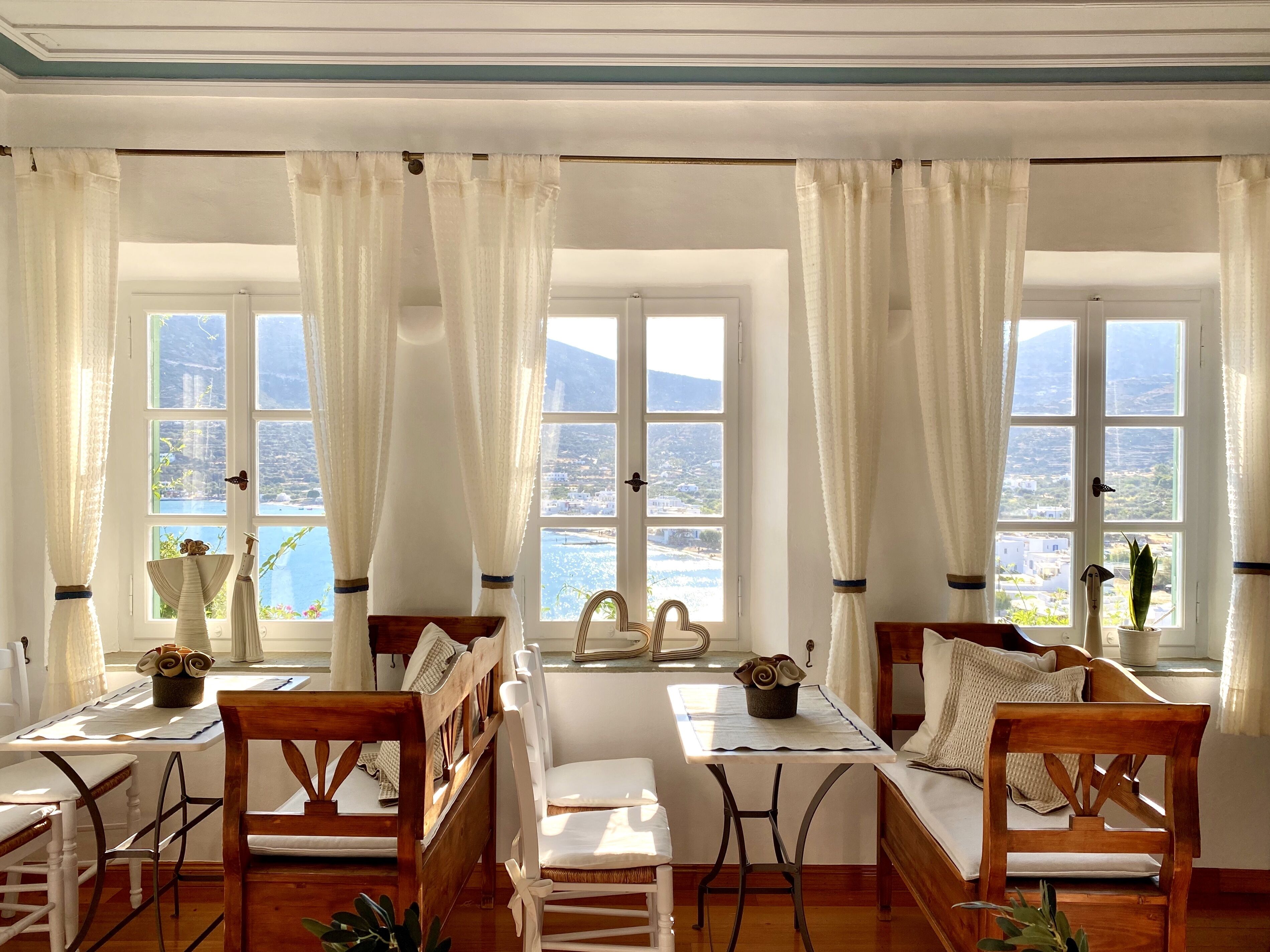 A room with a wooden table, two chairs, a sofa, and large windows with white curtains.