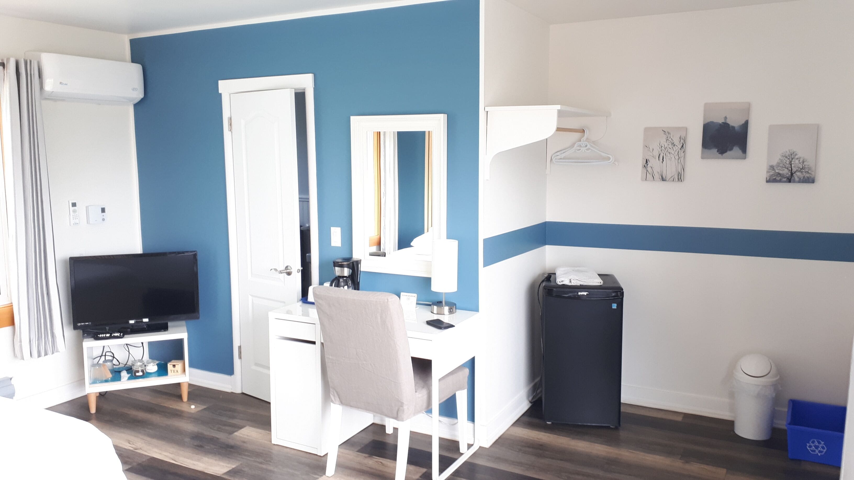 Habitació d'hotel amb una paret blava decorativa, un escriptori blanc amb una cadira, un televisor de pantalla plana, un mirall, un petit paper basc negre i un contenidor de reciclatge.