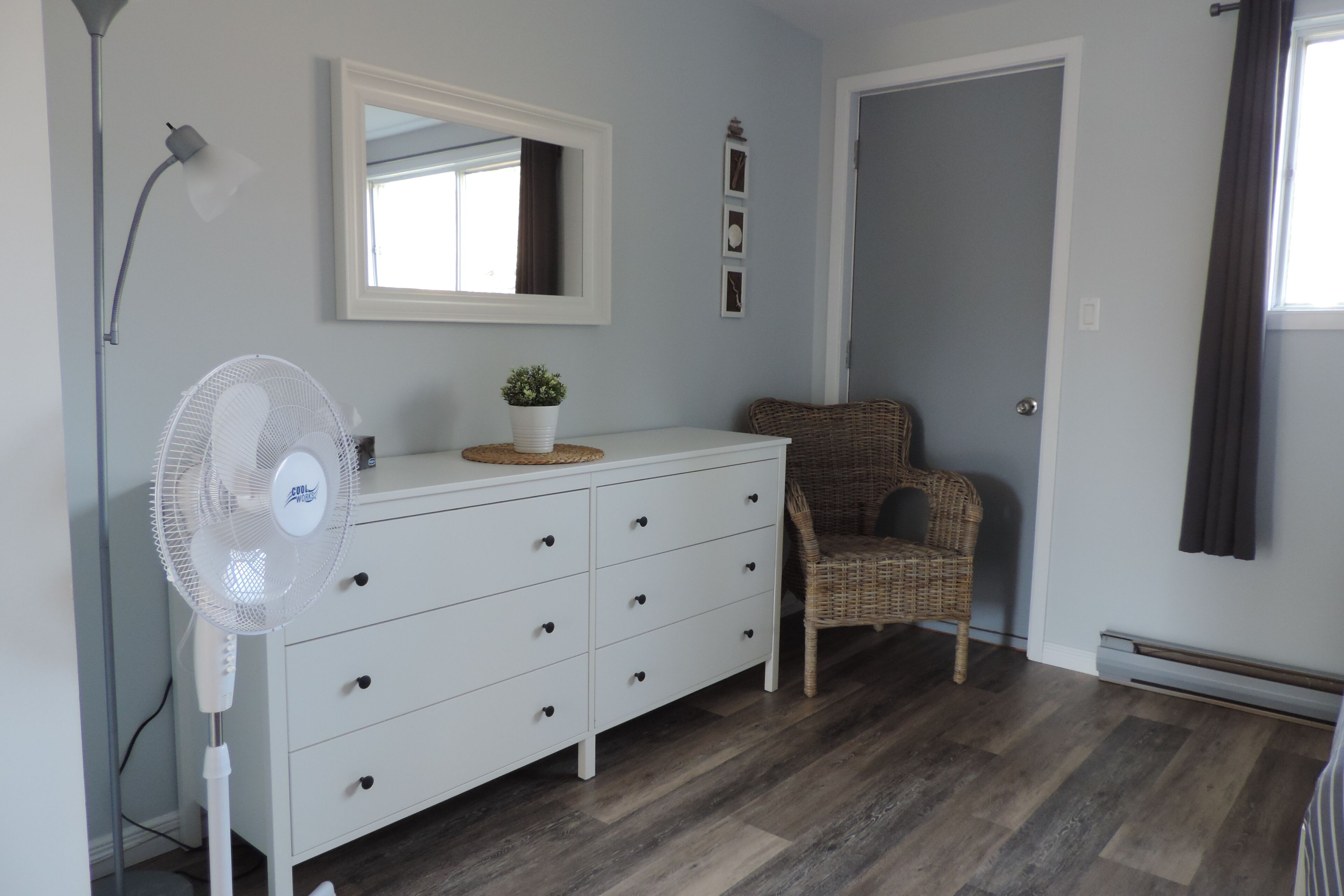 A room with a white dresser, a wicker chair, a standing fan, and a mirror on the wall.