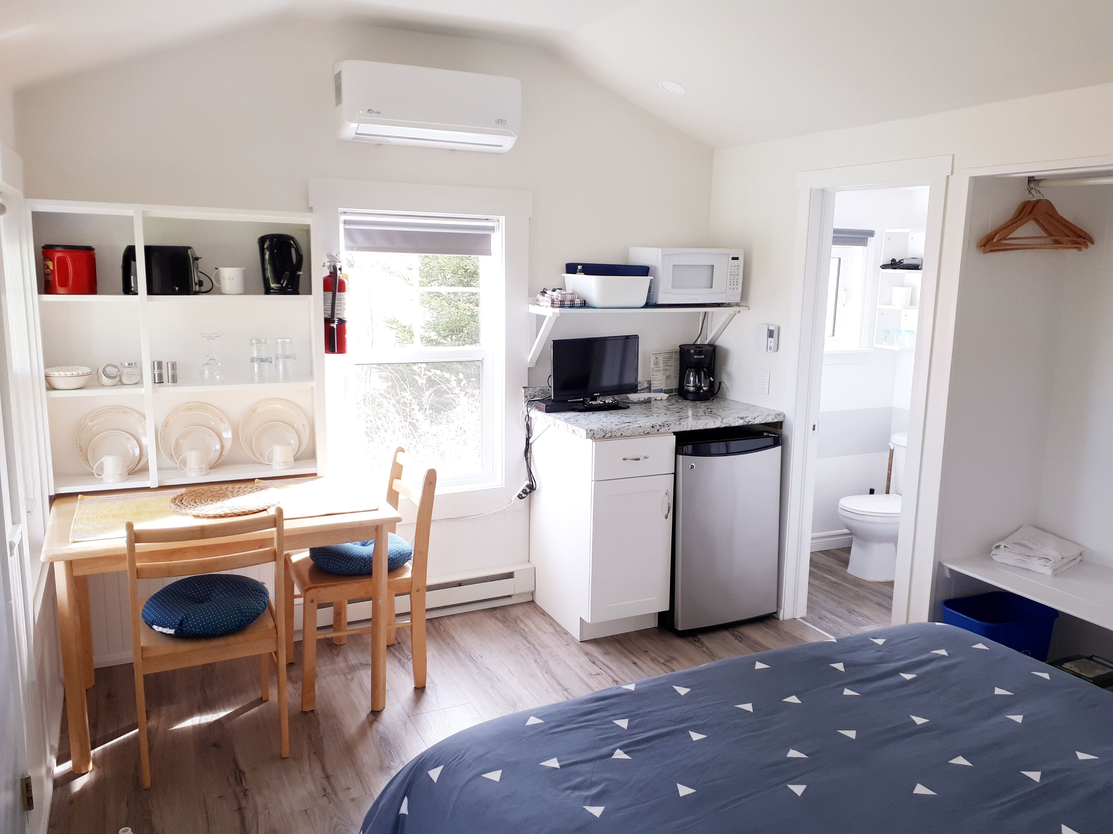 A compact, well-lit room with a kitchenette, a dining area with a table and chairs, a bed with a patterned bedspread, and a view of the outdoors through a window.