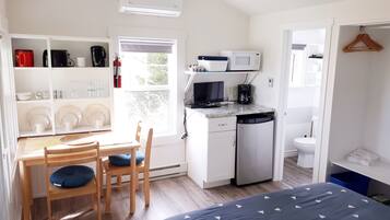 A compact, well-lit room with a kitchenette, a dining area with a table and chairs, a bed with a patterned bedspread, and a view of the outdoors through a window.