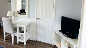 Una habitación con un escritorio blanco, una silla y un televisor. Hay una puerta y un espejo en la pared.