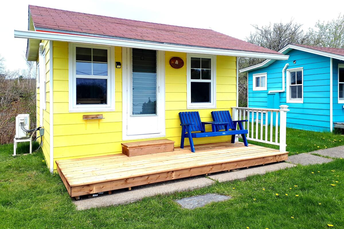 A yellow house with a red roof, a porch with two blue Adirondack chairs, and a blue house in the background.