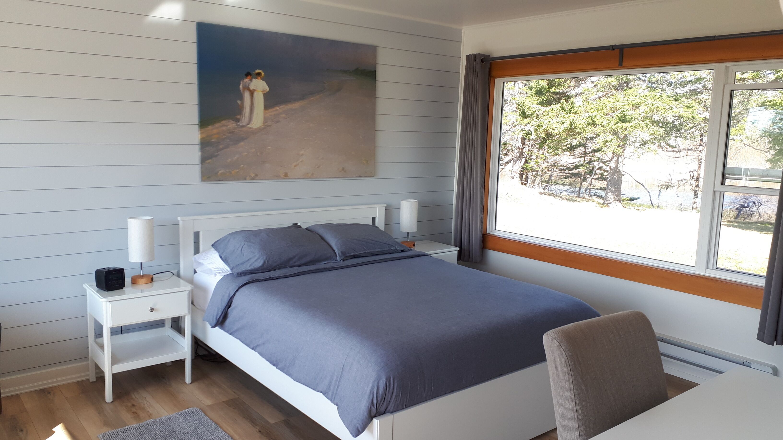 Un dormitorio con cama, mesitas de noche, una lámpara, un cuadro y una ventana con vista a árboles.