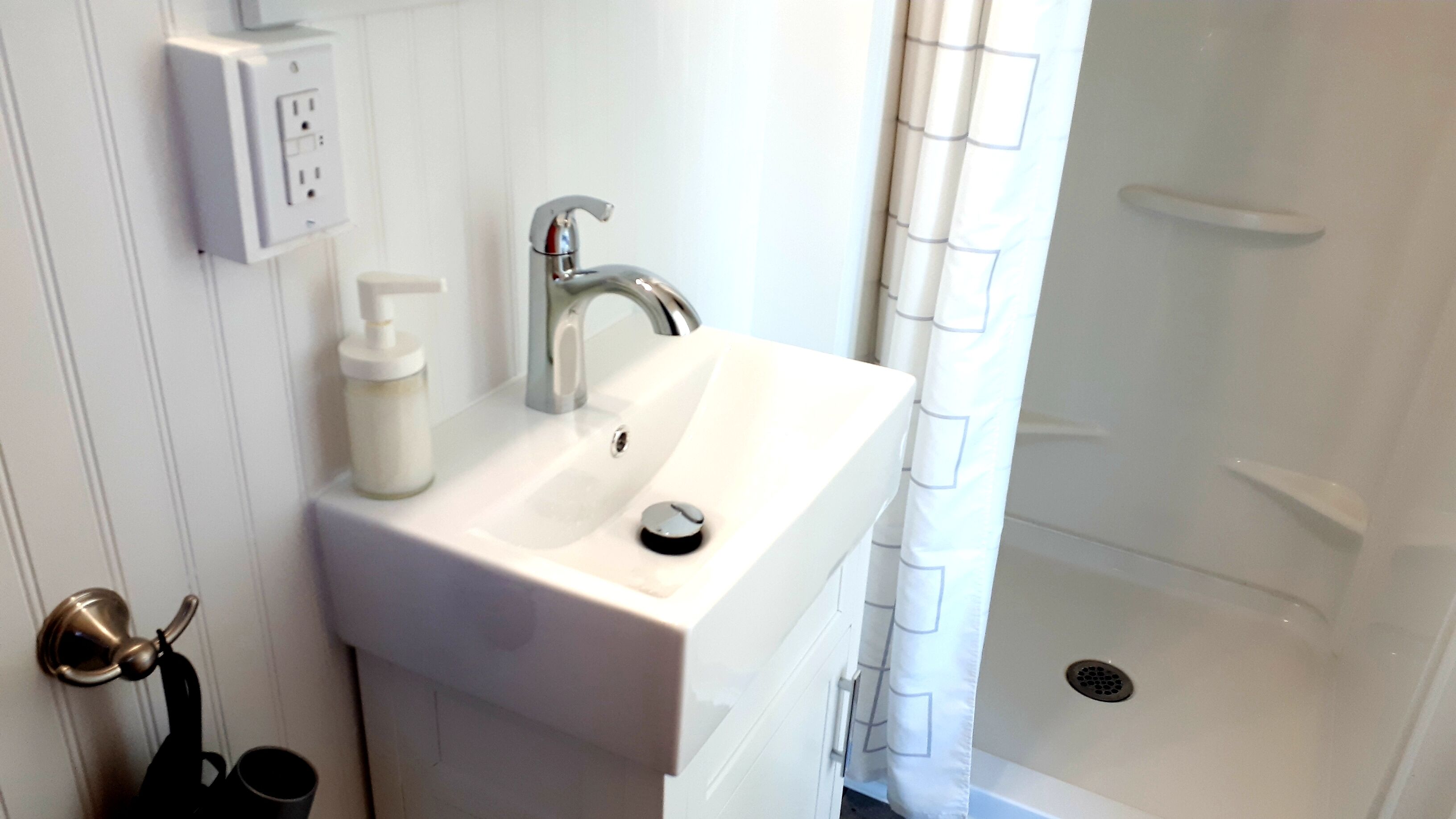 Un baño con lavabo, grifo y ducha blancos. Hay una botella de jabón y una taza negra sobre el lavabo.