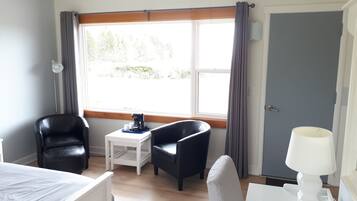 A room with a bed, two chairs, a small table with a coffee maker, a lamp, and a window with curtains.