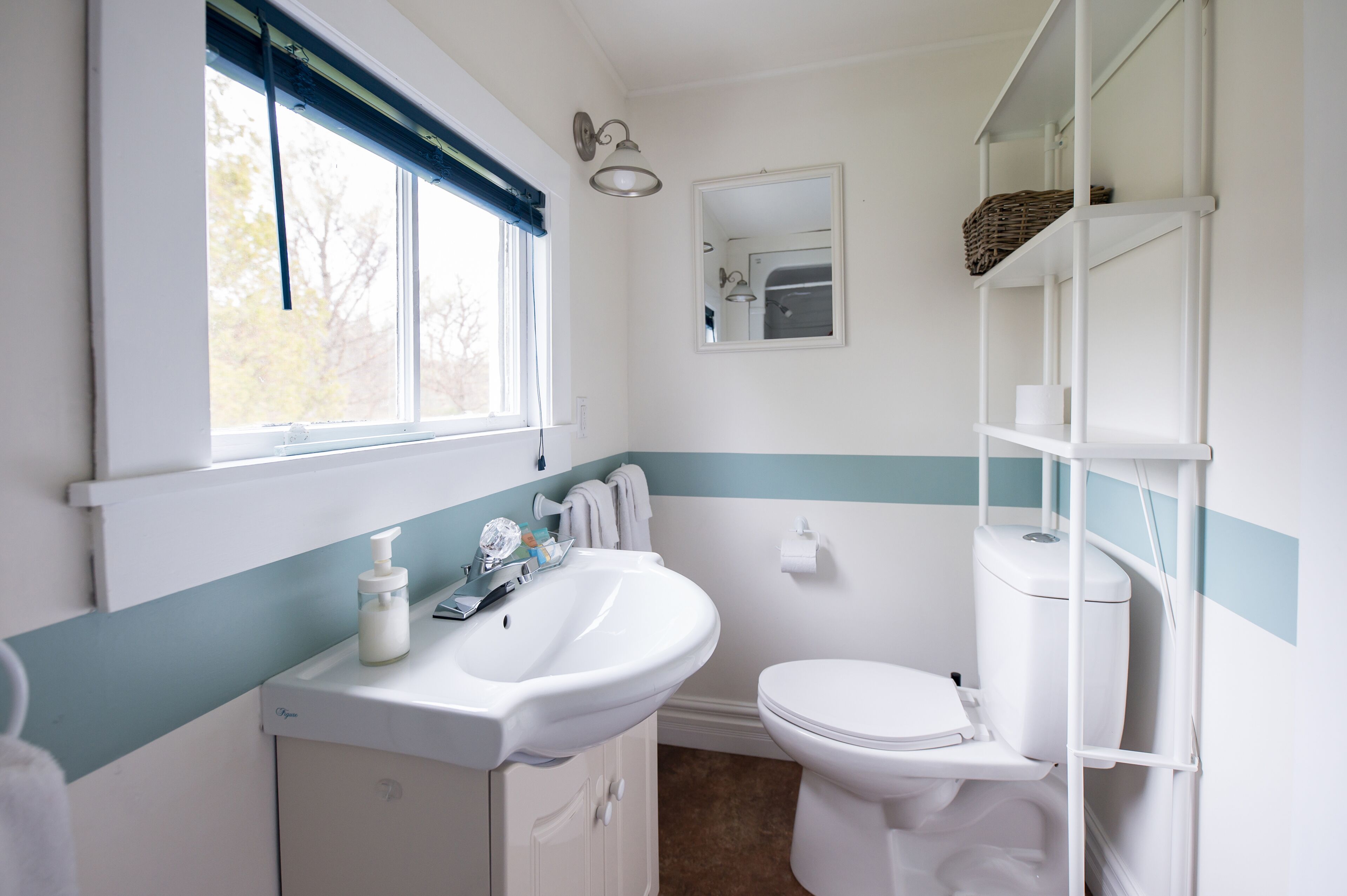 Een badkamer met een wit wastafel, toilet en raam met een blauw gordijn. Boven het wastafel hangt een spiegel en aan de muur een plank met een mandje.
