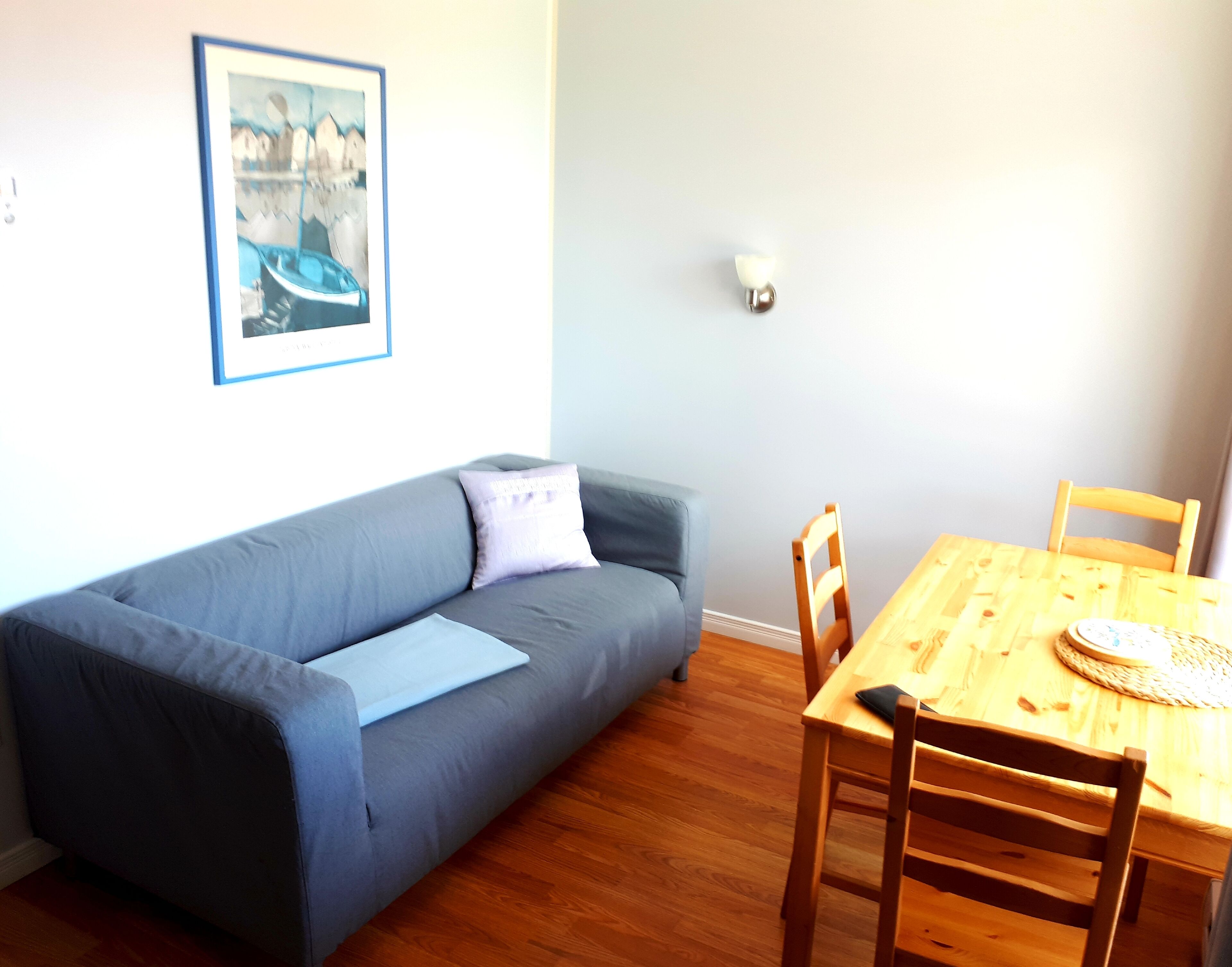 A living room with a grey sofa, a wooden dining table with chairs, a framed picture on the wall, and a small lamp.