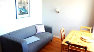 A living room with a grey sofa, a wooden dining table with chairs, a framed picture on the wall, and a small lamp.