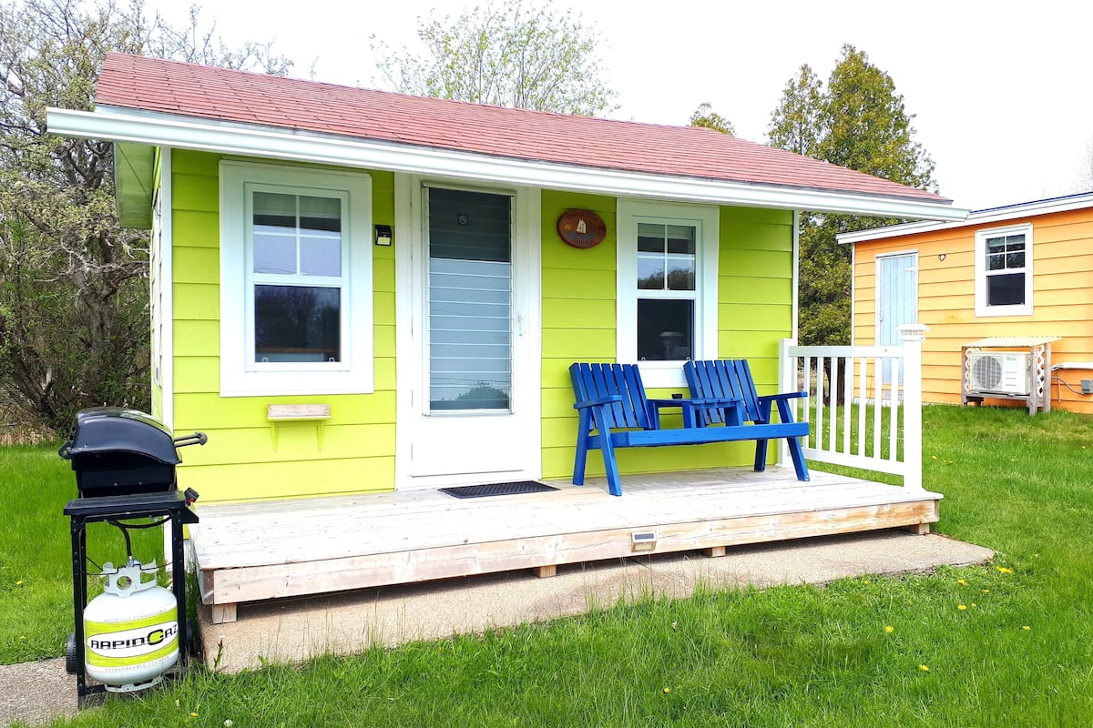 A green house with a red roof, a porch with blue Adirondack chairs, and a propane tank labeled "RapidCaz" on the side.