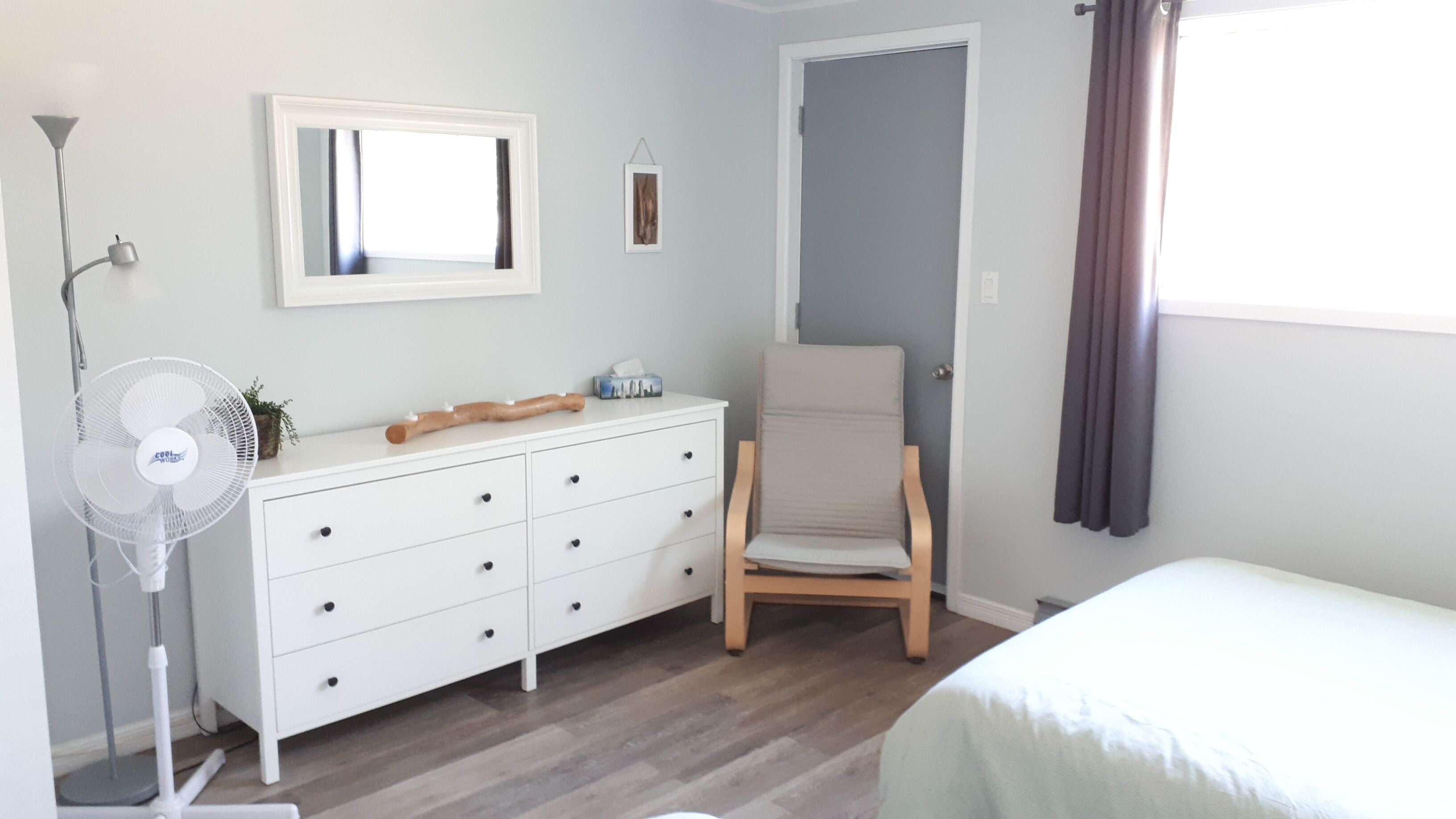 A bedroom with a white dresser, a chair, a bedside table, a standing fan, and a window with curtains.