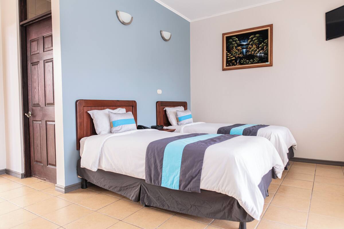 A hotel room with two beds, a wooden door, a wall-mounted TV, and a framed painting.