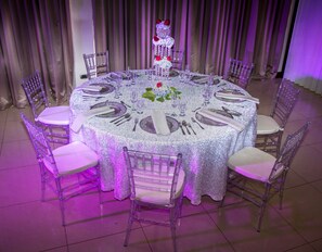 Een ronde tafel gedekt voor een formeel evenement, met transparante stoelen, een bloemrijk middelpunt en een paars verlichte achtergrond.