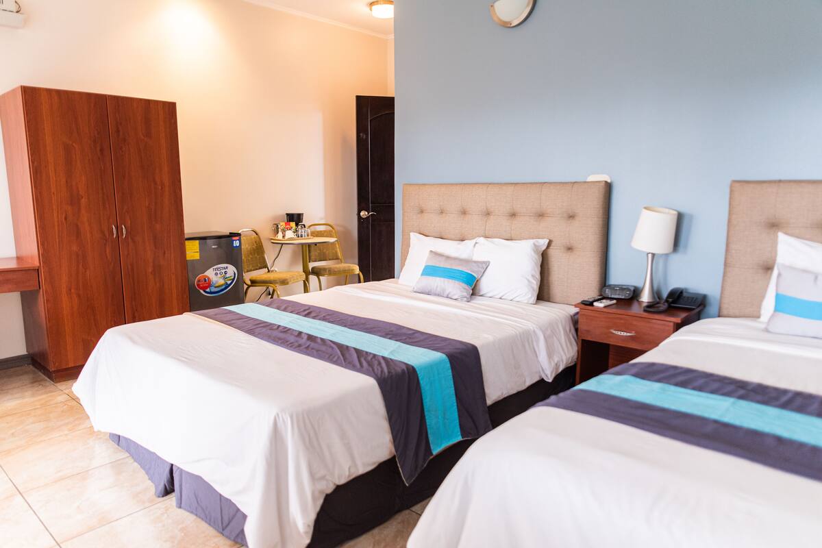 A hotel room with two beds, a wooden wardrobe, a small table with chairs, and a desk with a lamp and phone.