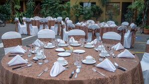 A round dining table set for a formal event, surrounded by chairs with white and brown sashes, in an outdoor setting with potted plants and a building in the background.