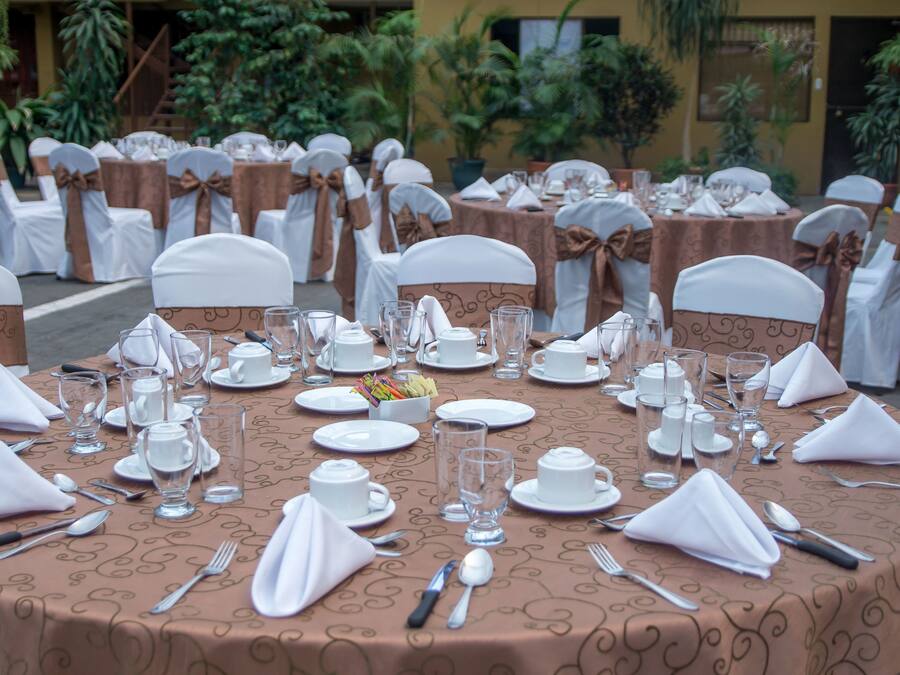 Una mesa redonda para comedor dispuesta para un evento formal, rodeada de sillas con cintas blancas y marrones, en un entorno al aire libre con plantas en macetas y un edificio al fondo.