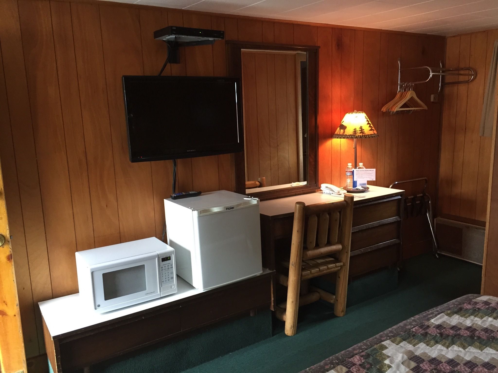 Una habitación con un revestimiento de madera en la pared, un televisor de pantalla plana sobre un refrigerador blanco, un escritorio de madera con una lámpara, una silla y una cama con colcha.