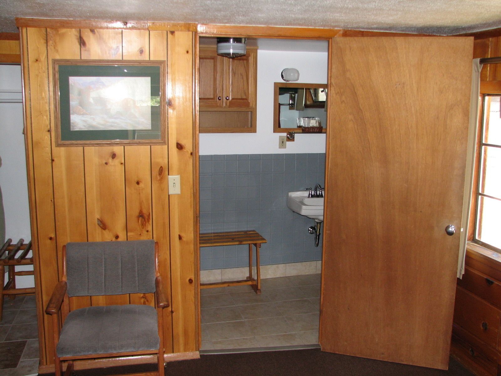A wooden-paneled room with a chair, a small table, and a door leading to a bathroom with a sink and mirror.