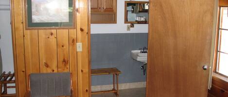 A wooden-paneled room with a chair, a small table, and a door leading to a bathroom with a sink and mirror.