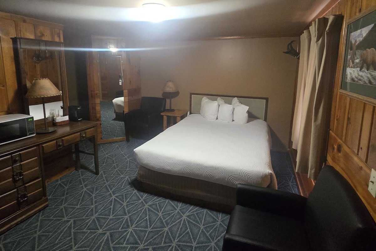 A hotel room with a large bed, a desk with a lamp, a chair, and a small table.