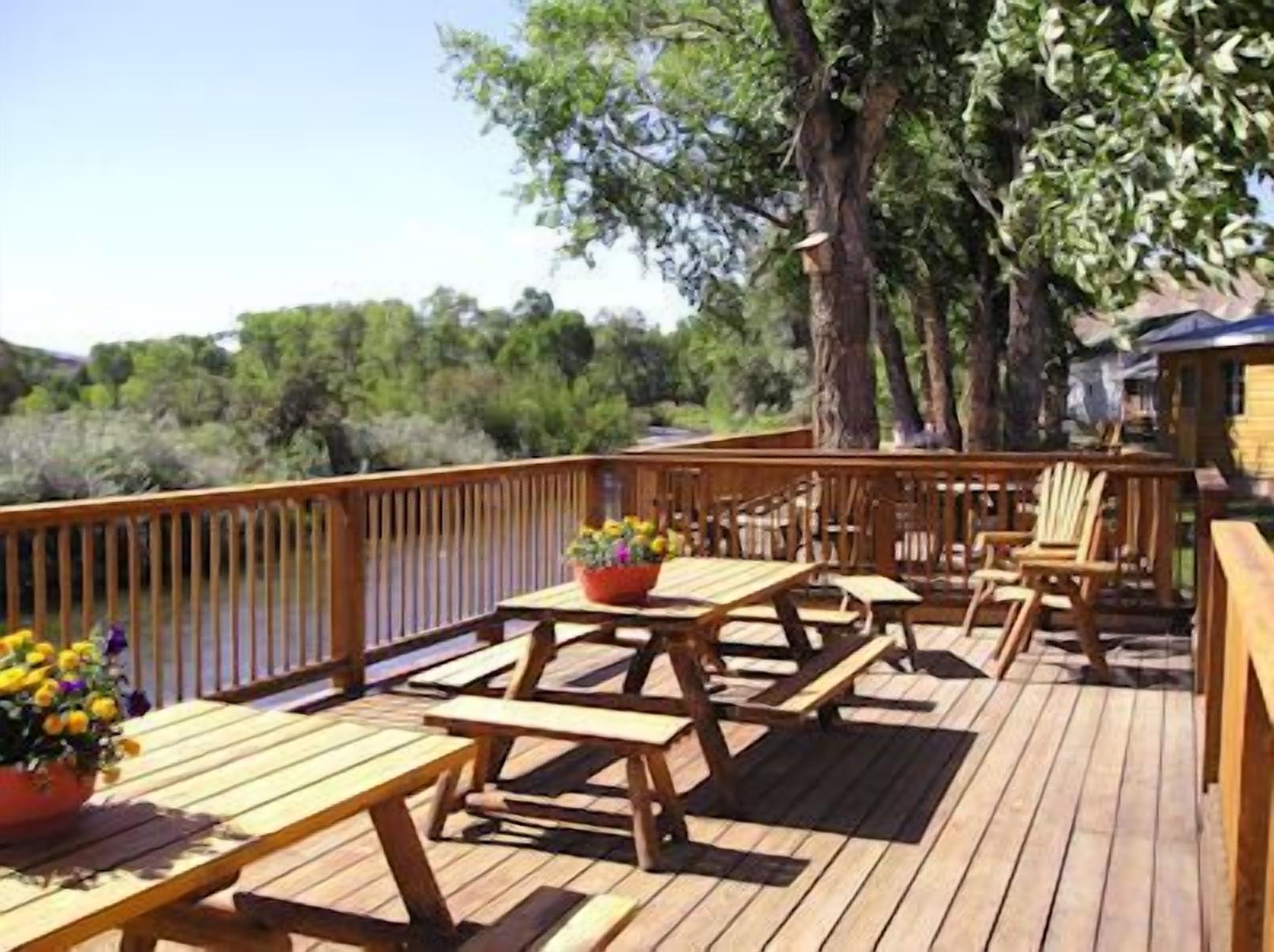 Eine Holzterrasse mit Picknicktischen und -stühlen, umgeben von Bäumen und Blick auf ein Gewässer.