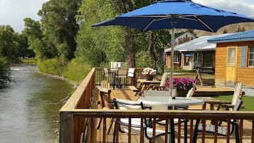 Una terraza de madera con mobiliario exterior, con vista a un río y un edificio de techo azul.