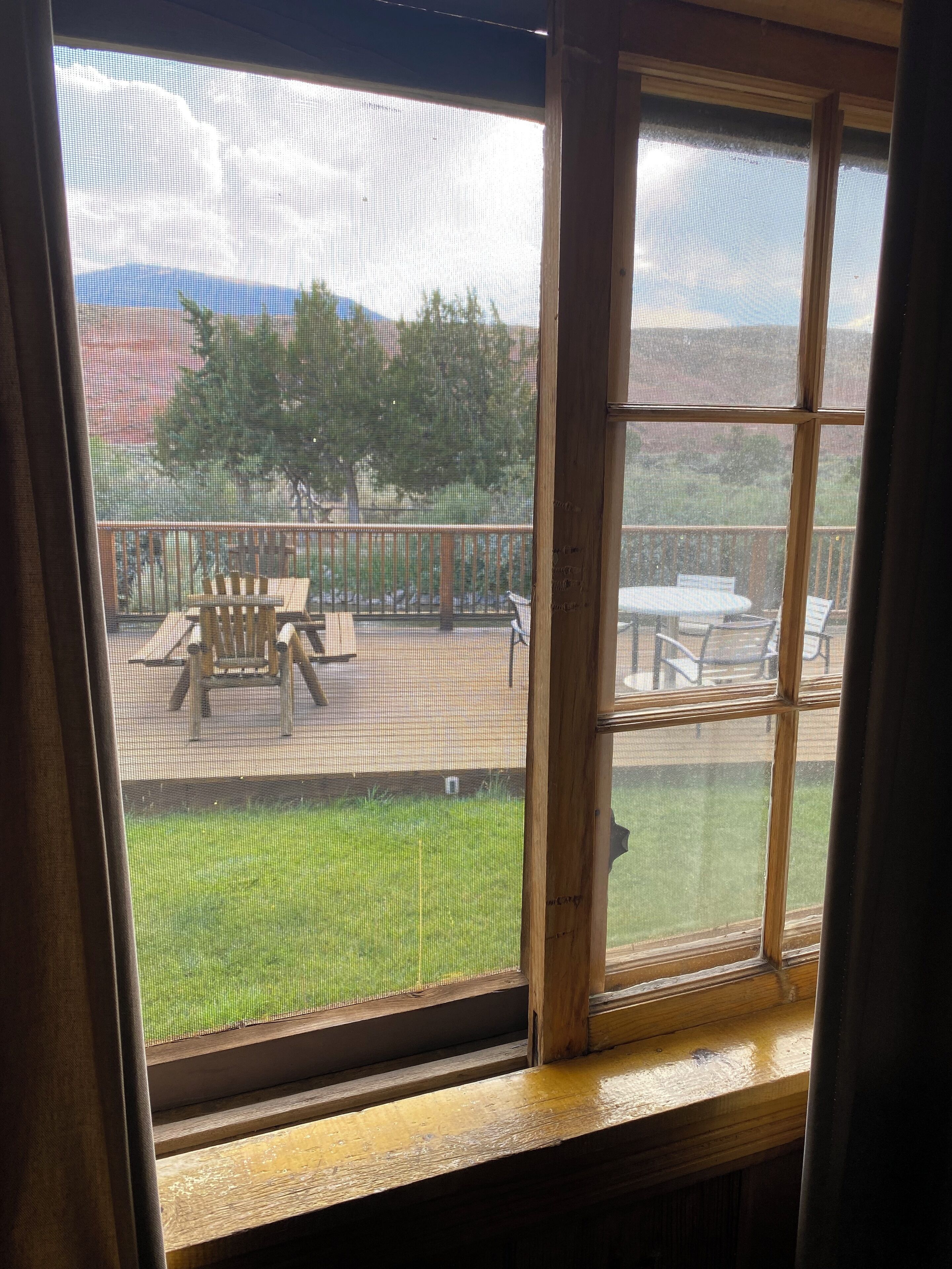 A view through a window with a wooden frame, looking out to a deck area with outdoor furniture and a lawn.