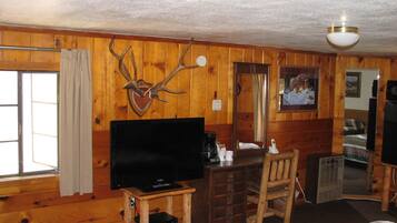 Ein Holzblockhaus-Innenraum mit einem Fernseher auf einem Holztischchen, einem Holzstuhl, einem kleinen Tisch, einem Spiegel und einem Kamin.