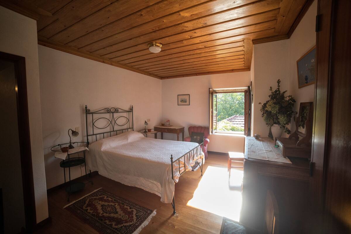 A bedroom with a wooden ceiling, a bed with a white bedspread, a small table with a lamp, a chair, and a window with a view of greenery.