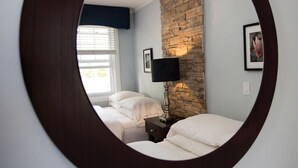 A circular mirror reflecting a bedroom with two beds, a stone accent wall, a bedside table with a lamp, and a window with blinds. - Studiotel Bromont (Bromont)