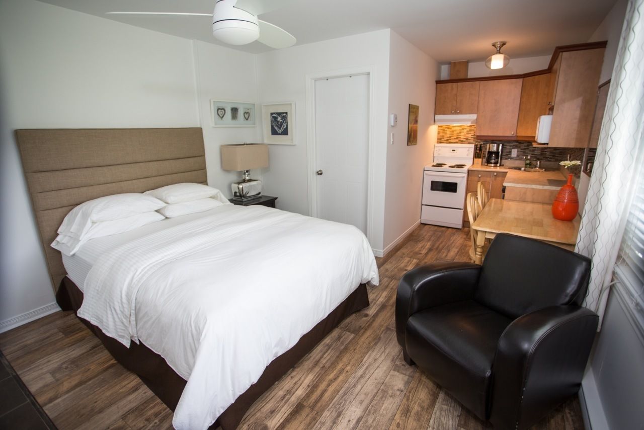 Een slaapkamer met een groot bed, een nachtkastje met een lamp en een stoel. Grenzend daaraan een kitchenette met een kookplaat, oven en koffiezetapparaat.