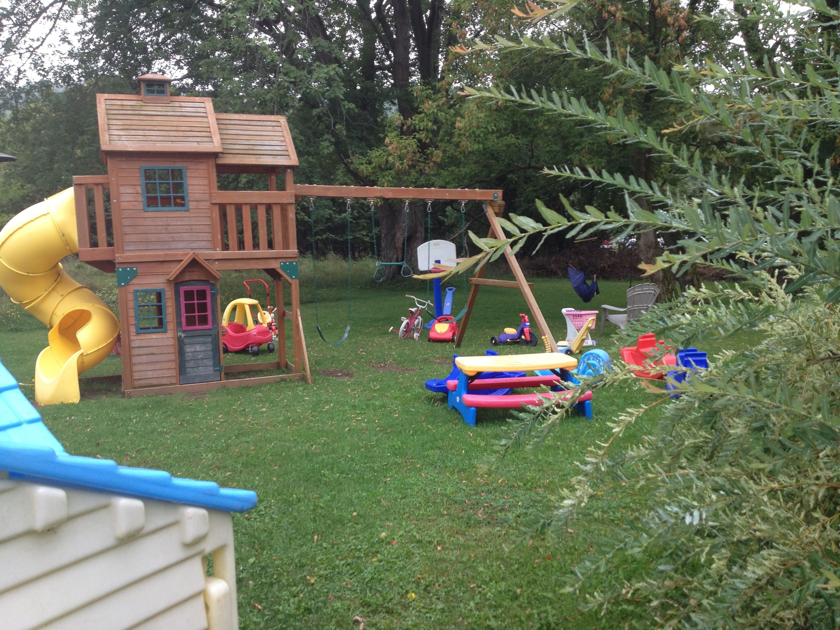 A wooden playhouse with a slide, a swing set, and various toys scattered around.