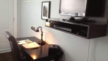 A small, dark wood desk with a chair, a flat-screen TV mounted on the wall, a small shelf below the TV, and a desk lamp.