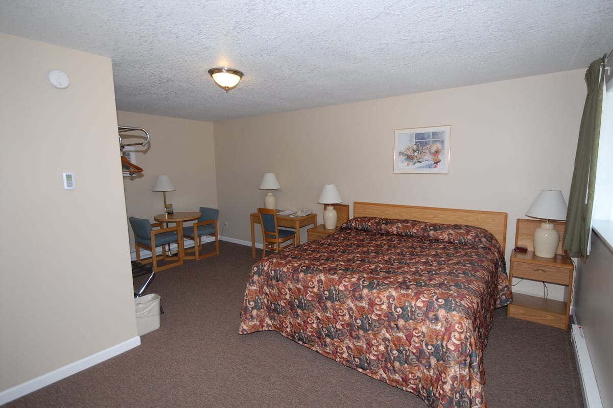 A hotel room with a bed, a desk, two chairs, a lamp, a painting, and a ceiling light.