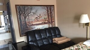 Una habitación de hotel con un sofá de cuero negro, una colcha de cama con motivos florales, un cuadro de pájaros sobre un puente y una lámpara de noche.