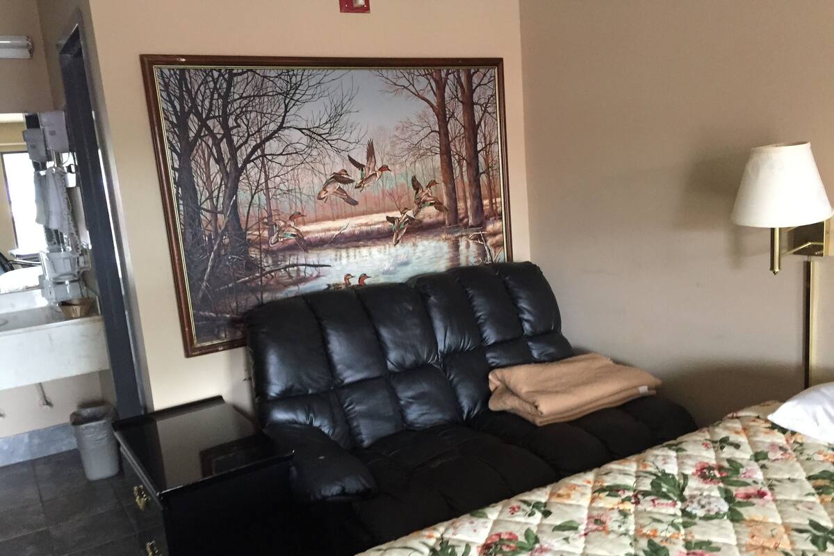 A hotel room with a black leather sofa, a floral bedspread, a painting of birds on a bridge, and a bedside lamp.