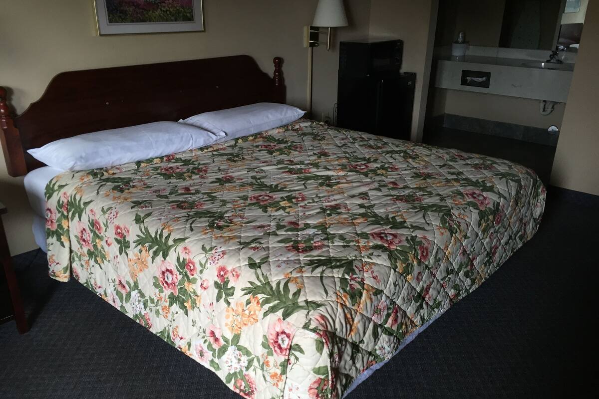 A hotel room with a bed, a nightstand, a lamp, a refrigerator, and a bathroom with a sink and mirror.