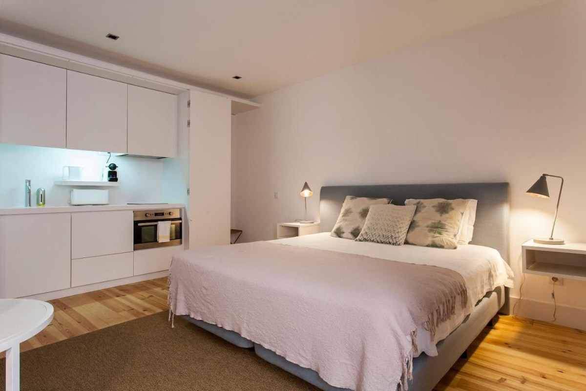 A bedroom with a bed, bedside tables, a nightstand, a desk, and a kitchenette.